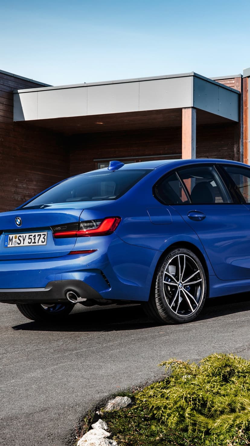 Blue BMW sedan parked on asphalt in front of a modern wooden building under a clear blue sky in bright daylight.