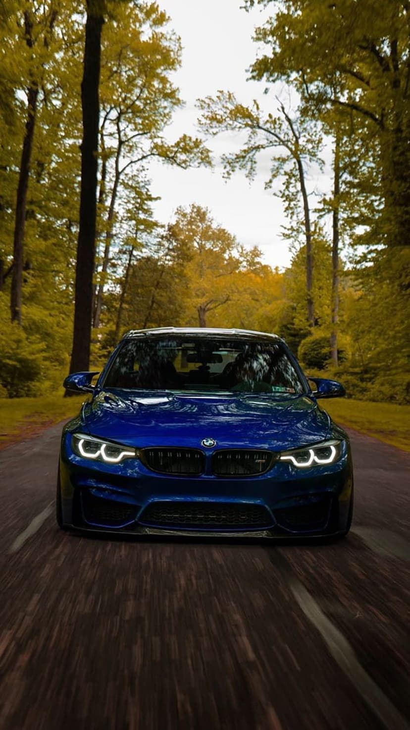 Blue BMW M4 sports car with glowing headlights driving on a forest road surrounded by yellow autumn leaves.