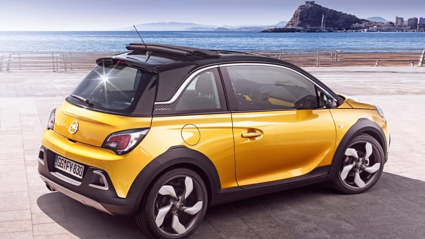 Yellow Opel Adam Rocks with an open roof parked by the blue sea and a coastal town under a bright sunny sky.