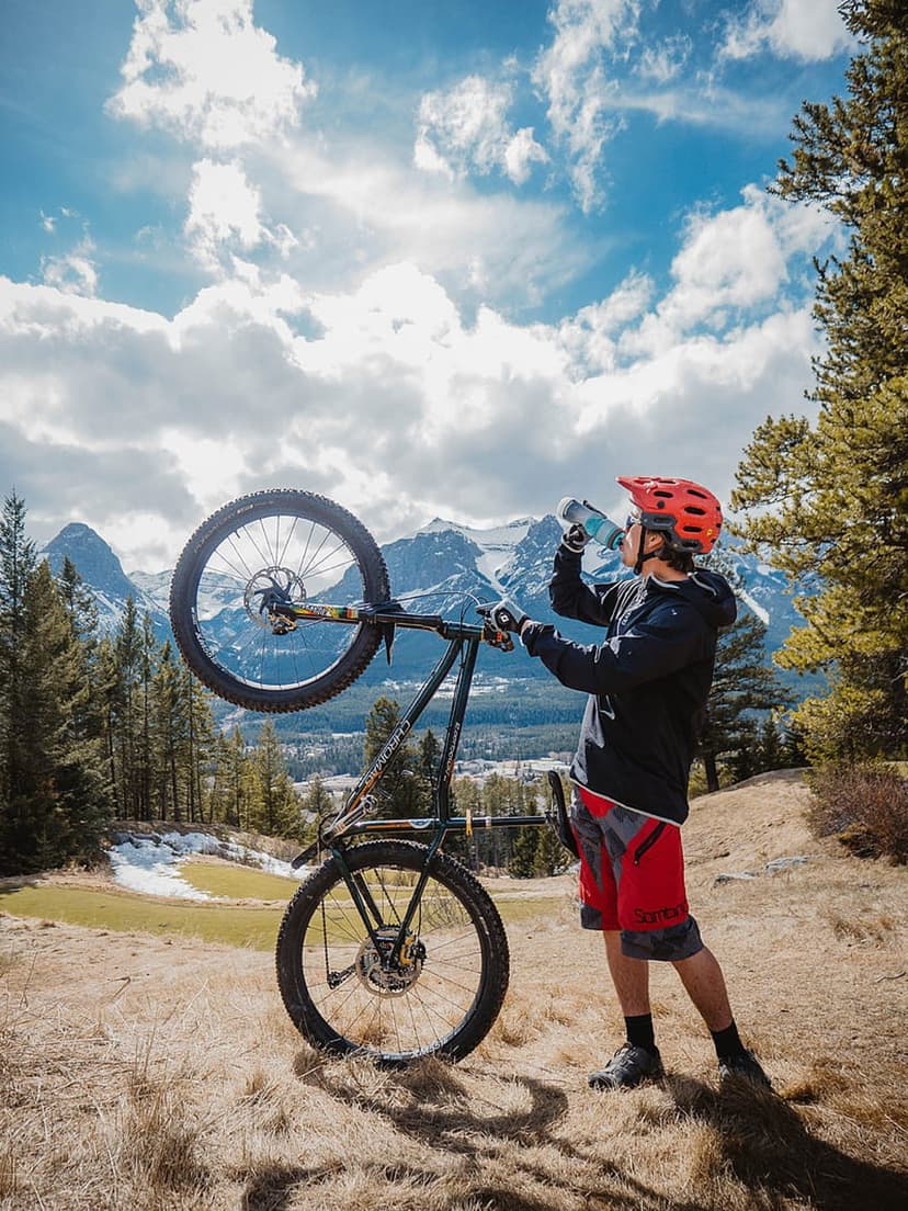 Male cyclist in red shorts drinks from a bottle beside his bike with snow-capped alpine mountains behind him.