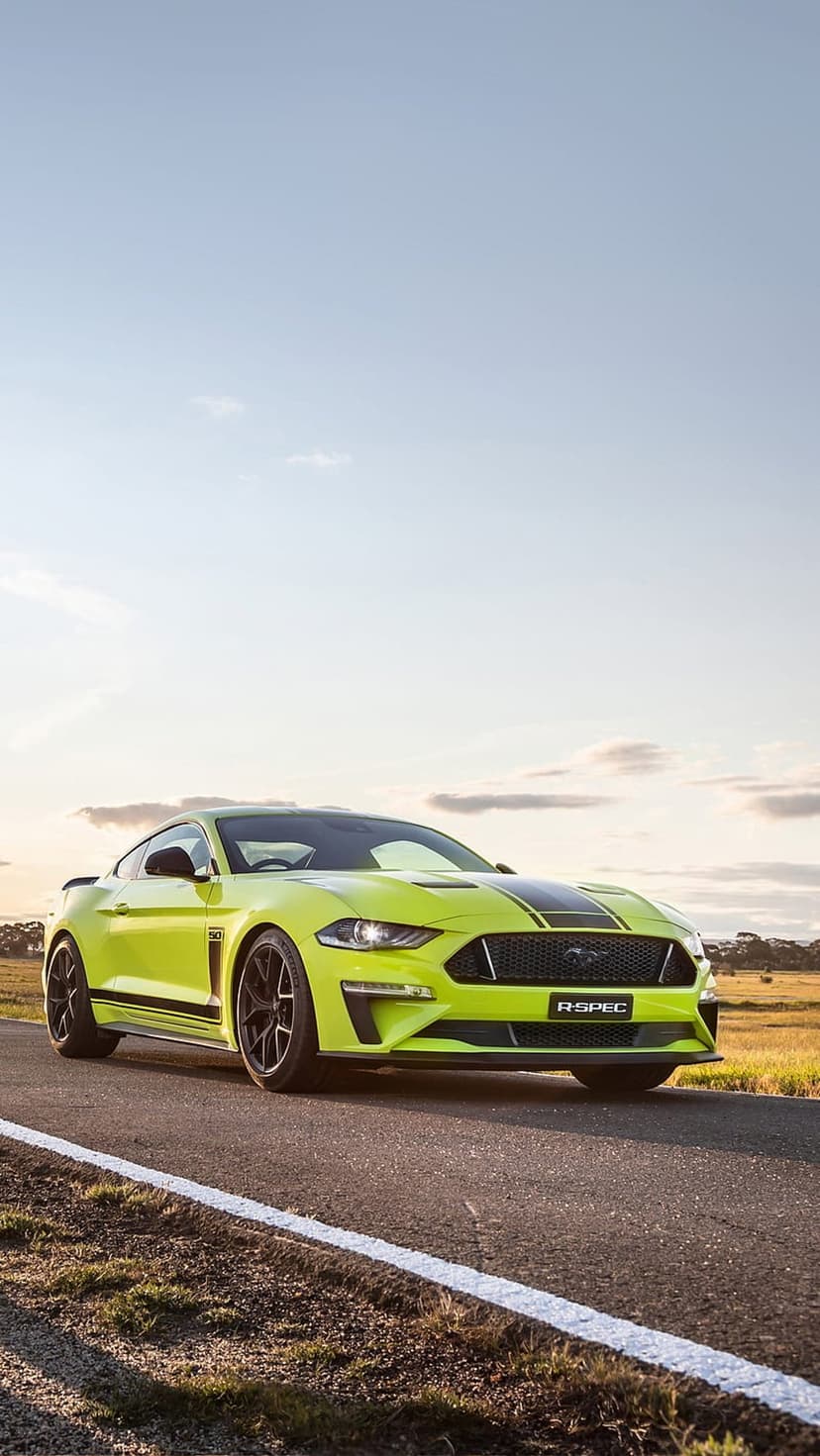 Lime green Ford Mustang R-Spec with black racing stripes cruises on a rural road during a warm golden hour.