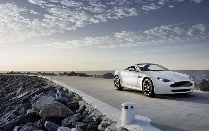 White Aston Martin convertible on a concrete coastal road next to rocks and the ocean under a cloudy sky.