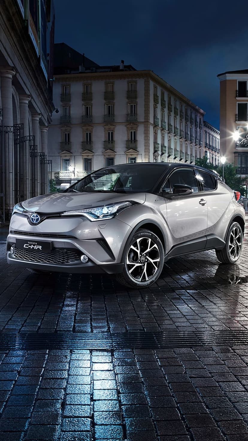 Silver Toyota C-HR parked on wet cobblestones at night with urban building reflections on the glossy paint.