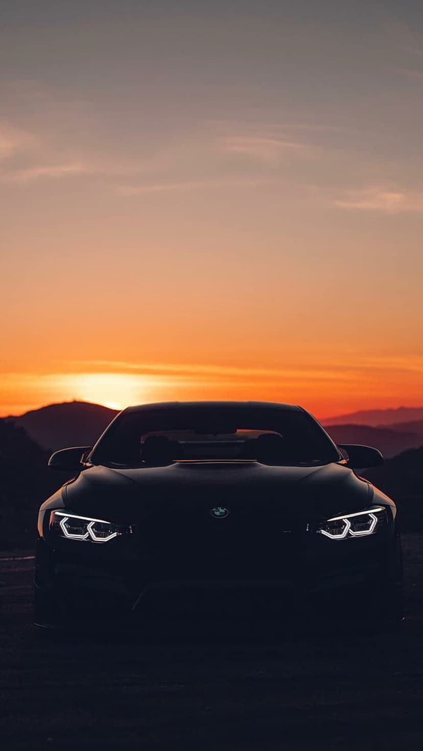 Black BMW convertible with glowing headlights parked on a mountain ridge during a purple and orange sunset.