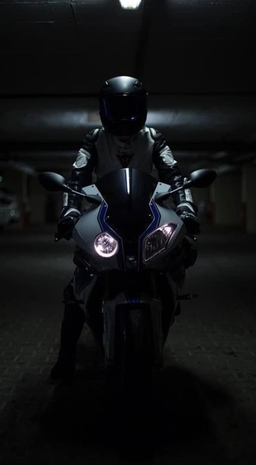 Motorcyclist in black gear sits on a sportbike with glowing headlights in a dark, shadowy garage interior.