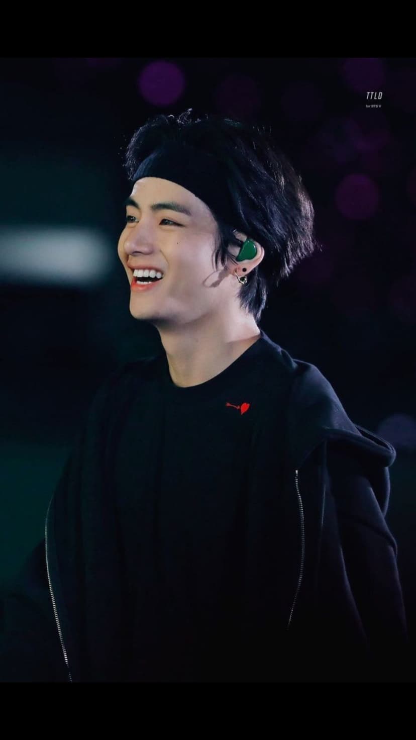 Kim Taehyung laughing in a black hoodie and headband with purple stage lights blurred in the background.