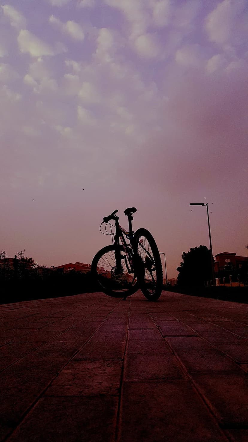 Bicycle silhouette on a textured paved path under a purple and pink sunset sky with scattered evening clouds.