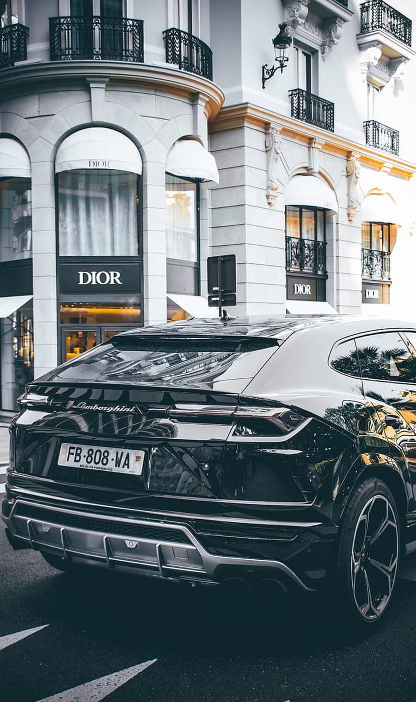 Black Lamborghini Urus SUV parked on a city street in front of a Dior boutique with arched stone windows.