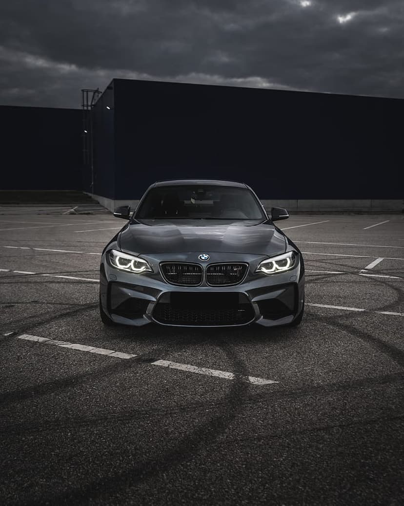 Dark gray BMW M2 coupe parked on asphalt with tire marks against a cloudy sky and industrial building backdrop.