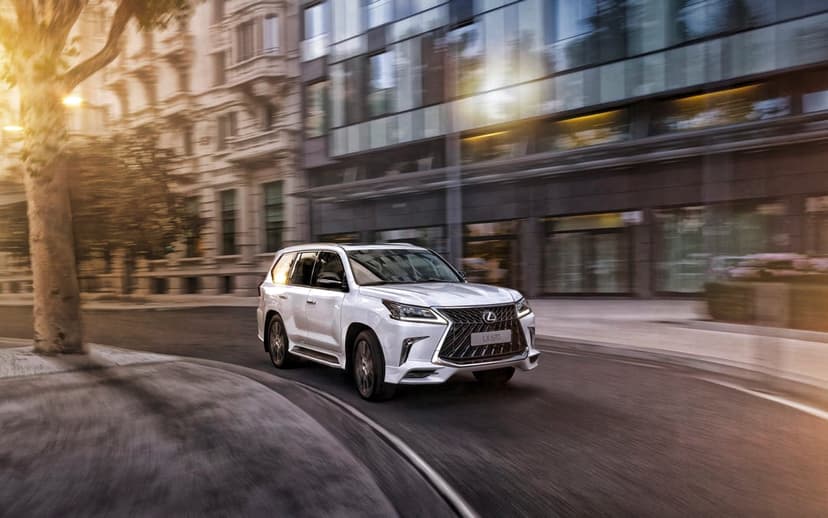 White Lexus LX 570 SUV driving on a city street with motion blur and warm golden hour sunlight reflections.