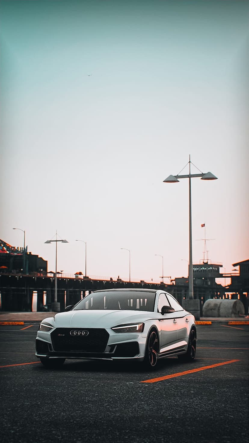 White Audi RS5 parked on a pier at sunset with a twilight sky and industrial structures in the background.