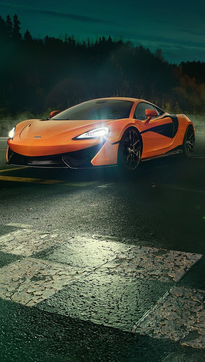 Orange McLaren hypercar with glowing headlights driving on a wet asphalt road through a dark forest at twilight.