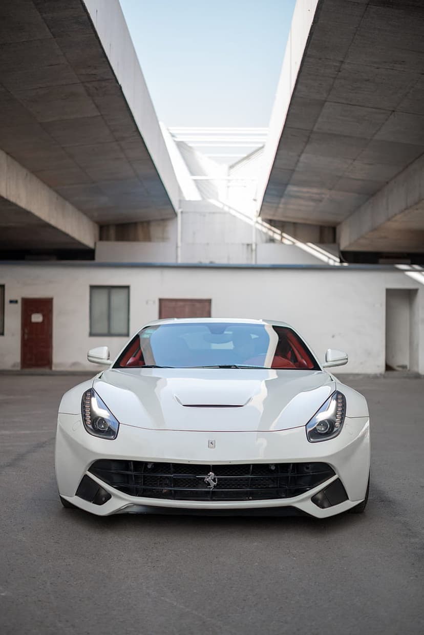 White Ferrari F12 parked on asphalt beneath a concrete brutalist overpass with sunbeams hitting the hood.