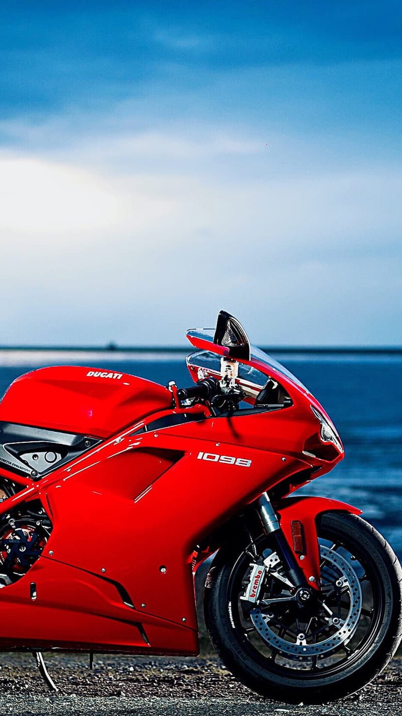 Red Ducati 1098 motorcycle parked on a sandy beach against blue ocean waves and a clear sunny horizon.