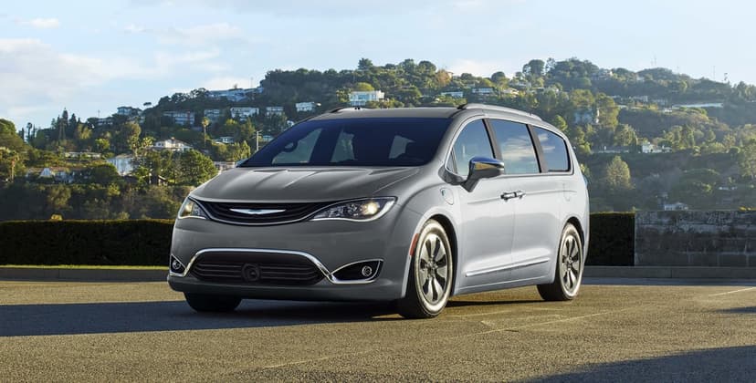 Silver Chrysler Pacifica Hybrid minivan parked on asphalt against a sunlit hillside with houses and trees.