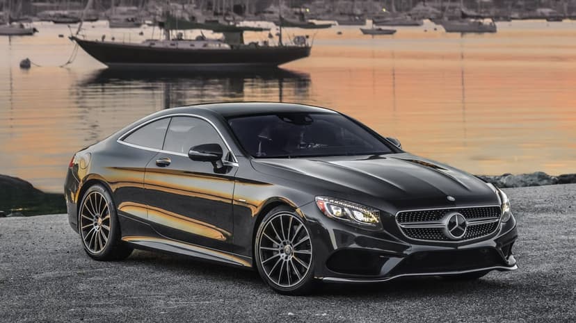 Charcoal Mercedes-Benz S-Class Coupe parked on a gravel pier beside a harbor with sailboats at sunset.