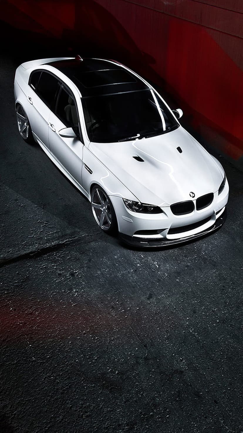 White BMW M3 sedan with a black roof and silver wheels parked on asphalt against a vibrant red textured wall.