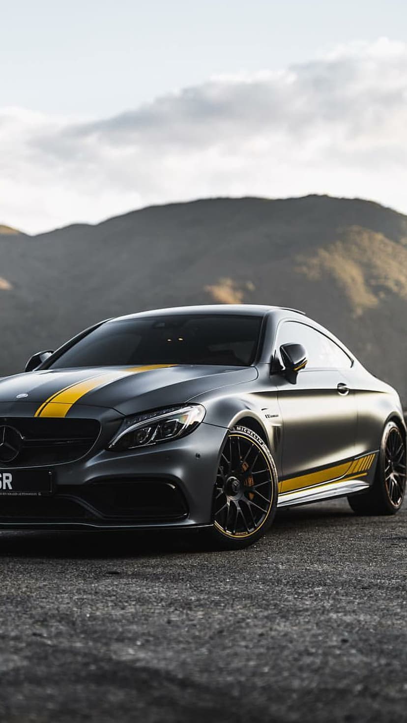 Mercedes-AMG C63 S Coupe with yellow stripes parked on asphalt before a backdrop of cloudy mountain peaks.