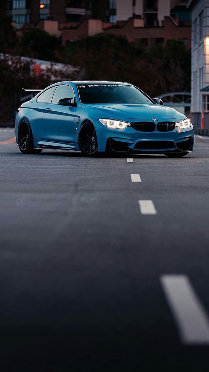Blue BMW M4 Coupe with a rear spoiler and glowing headlights parked on an asphalt road with white lane markings.