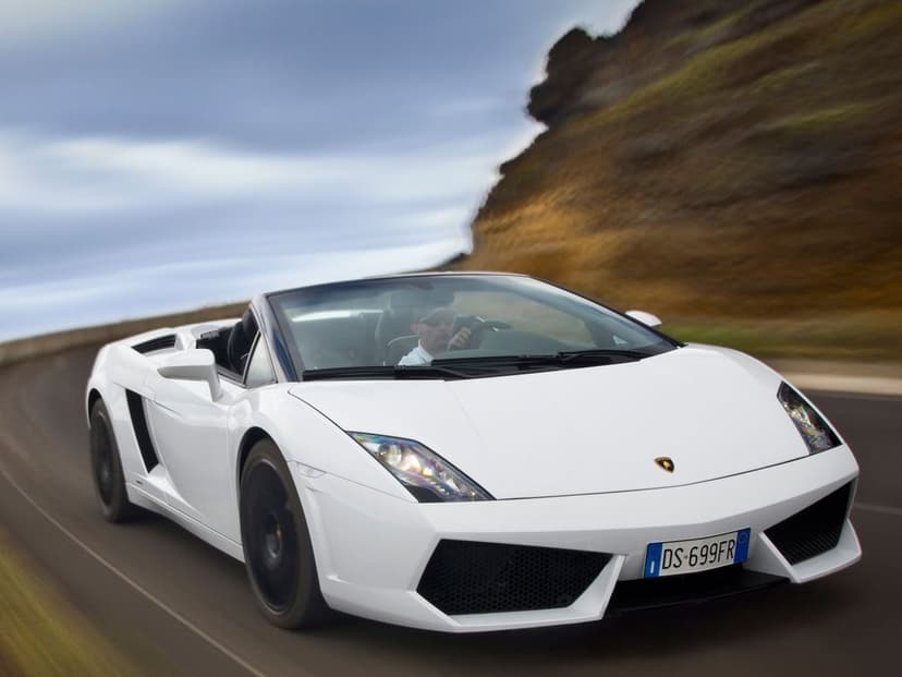 White convertible Lamborghini driving on a winding road next to a rocky cliffside under a moody cloudy sky.