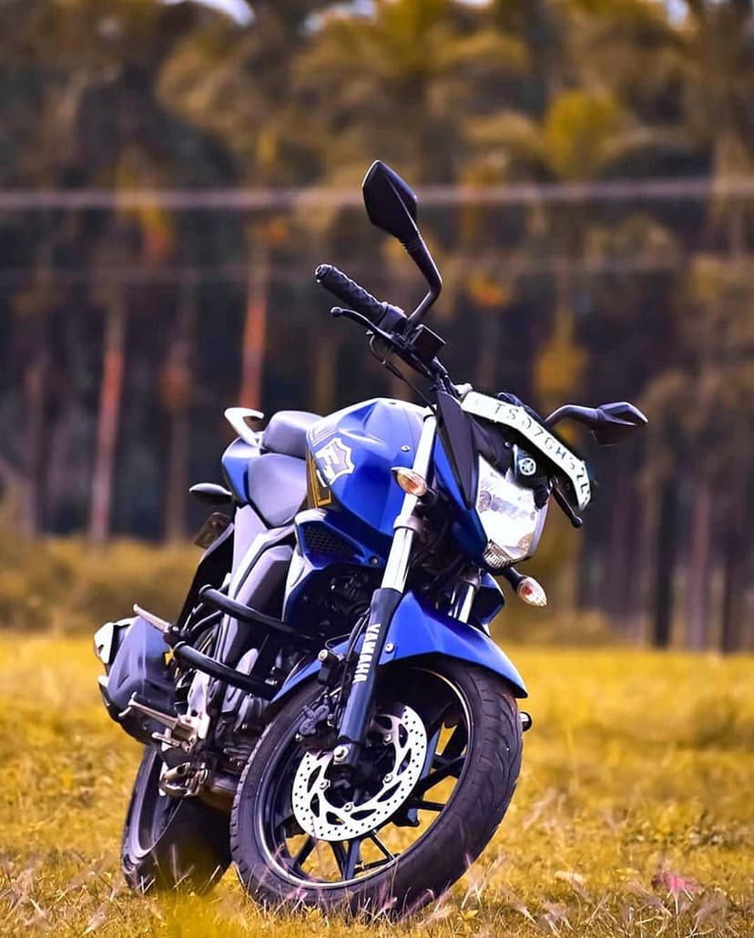 Blue Yamaha sportbike with black wheels parked in a sun-drenched grassy field against a blurred tree line.