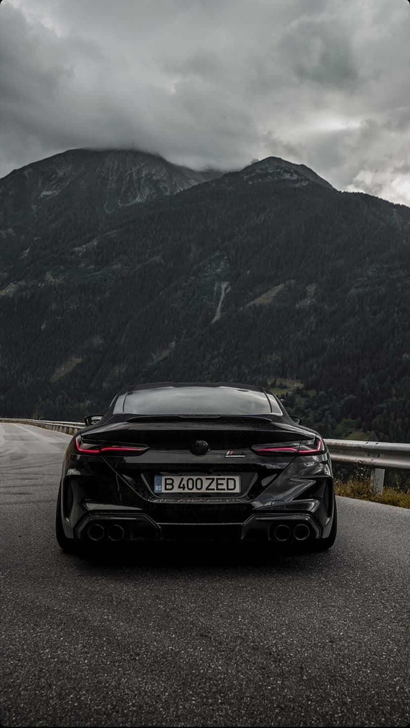 Black BMW 8 Series coupe parked on an asphalt mountain road with red taillights and dark forest trees.
