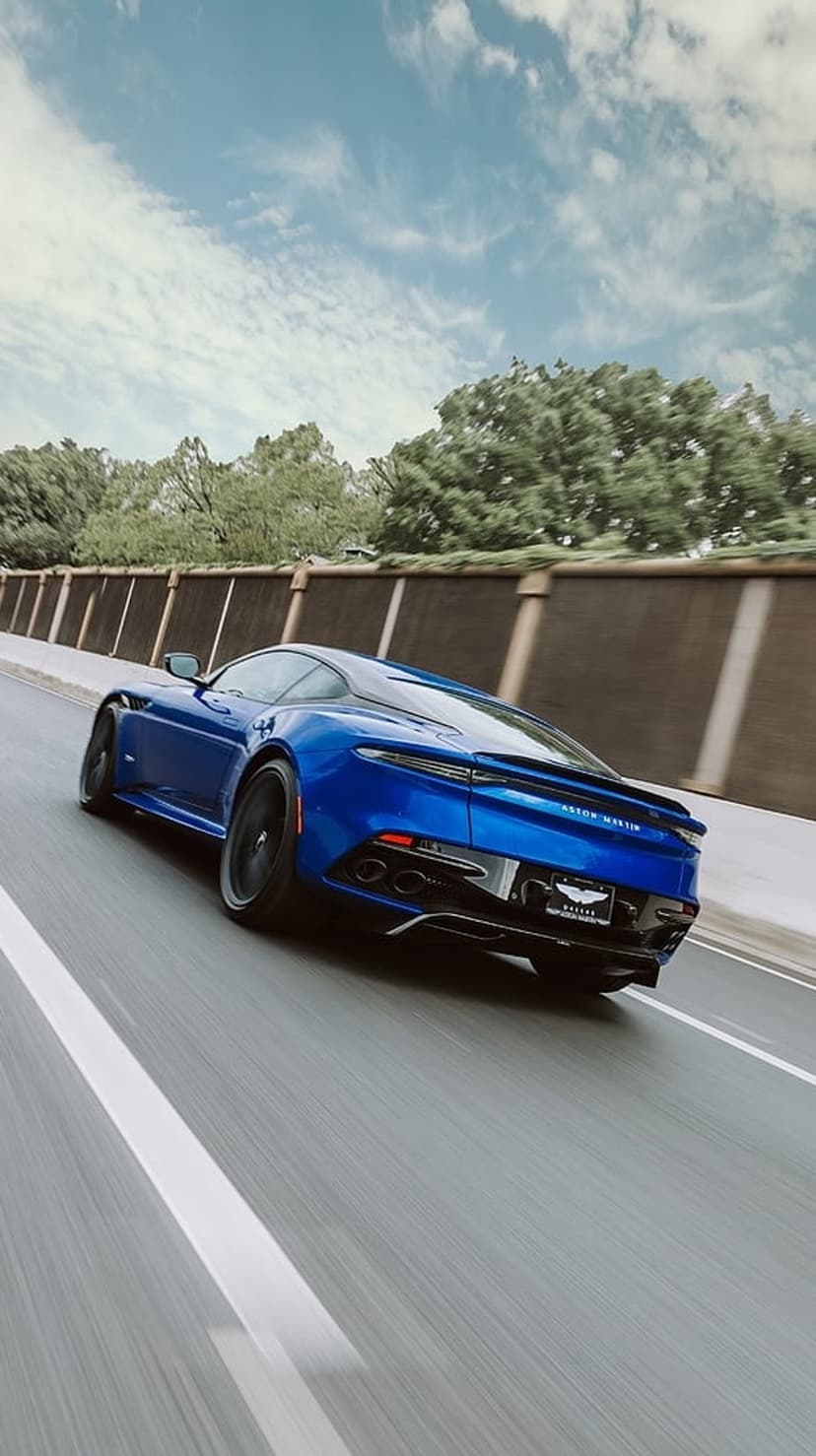 Blue Aston Martin DBS Superleggera sports car speeding on a sunlit road with motion-blurred green trees.