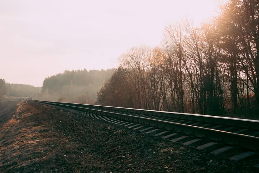 Iron railroad tracks stretch toward a misty autumn forest under a glowing orange golden hour sunset sky.