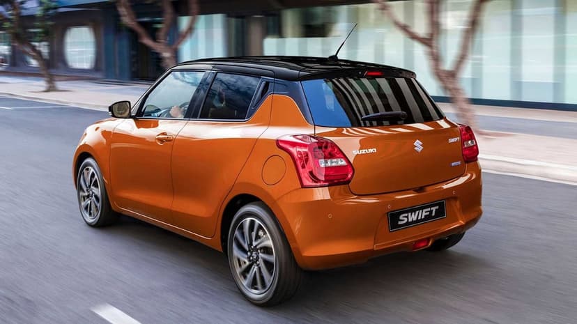 Metallic orange Suzuki Swift driving on a city road with a blurred urban background and black contrast roof.