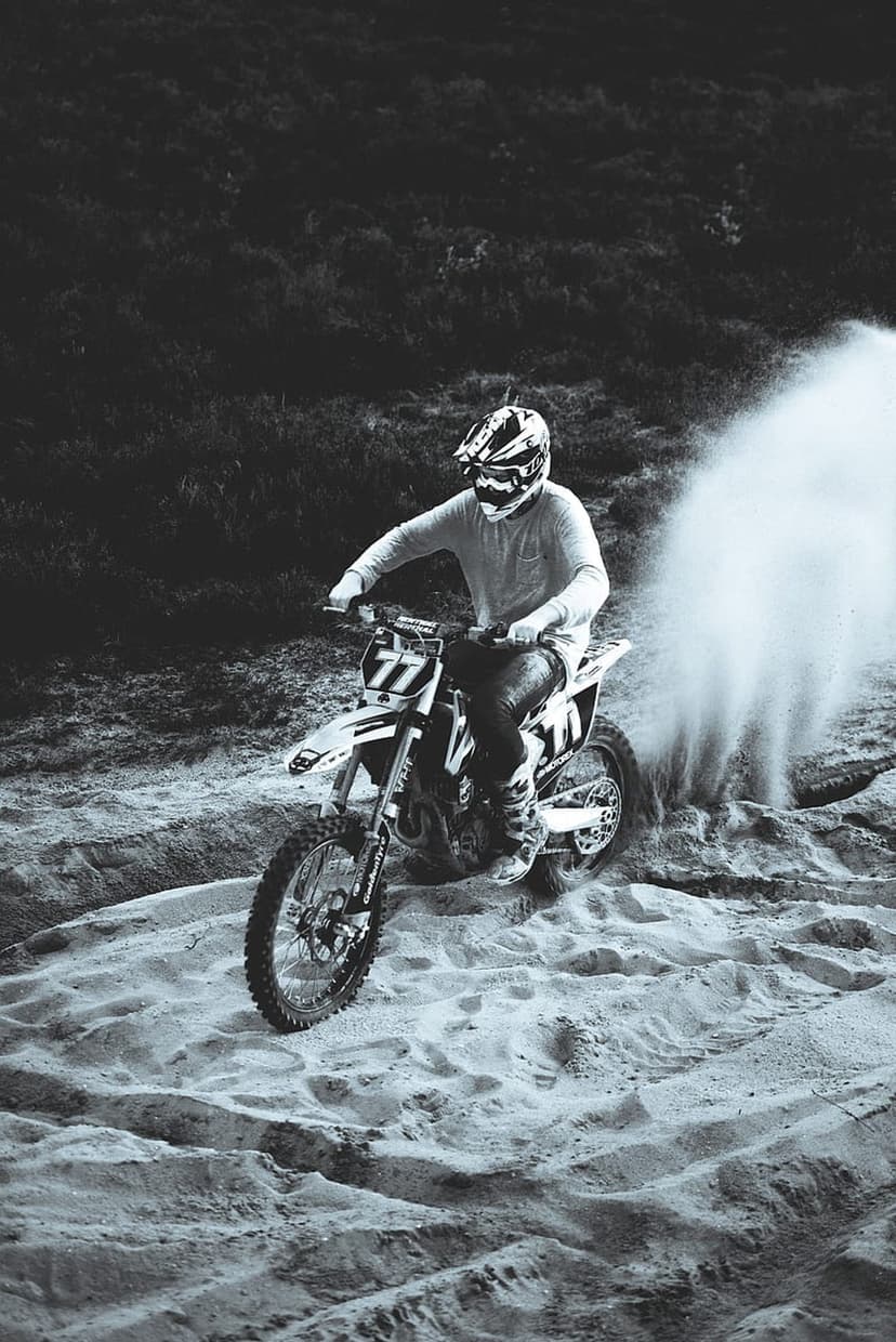 Motocross rider wearing a helmet leans a dirt bike into a sandy turn, kicking up a large plume of dust in monochrome.