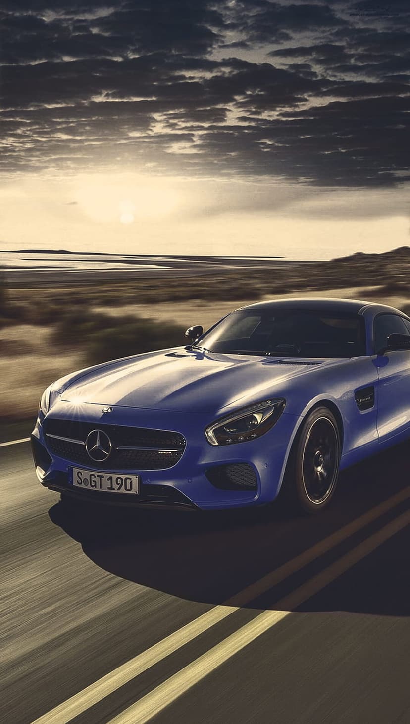 Blue Mercedes-AMG GT S sports car speeds on a road under a dramatic sunset sky with motion blur and golden light.