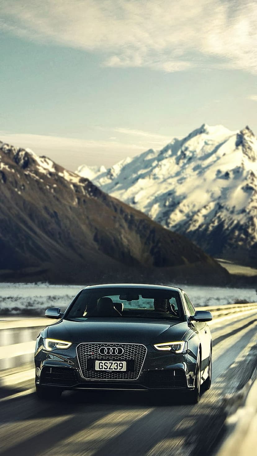 Audi car driving on a snowy mountain road with jagged peaks and frosted evergreen trees in the distance.
