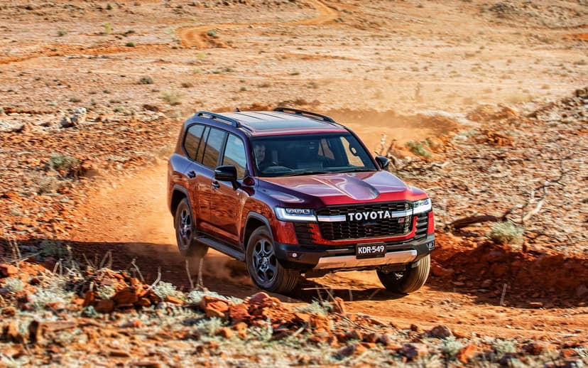 Red Toyota Land Cruiser SUV driving on a rocky desert track kicking up dust under bright daylight.