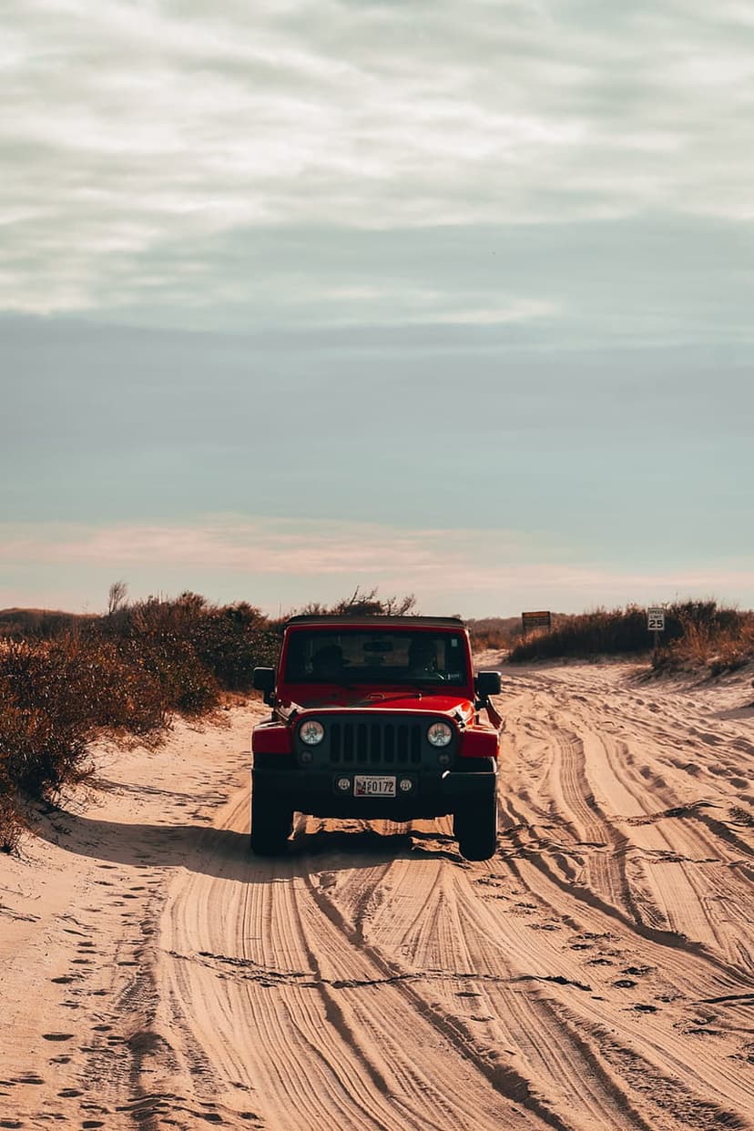 Red Jeep Wrangler drives through sandy dunes kicking up dust trails under a cloudy sky during an off-road trip.