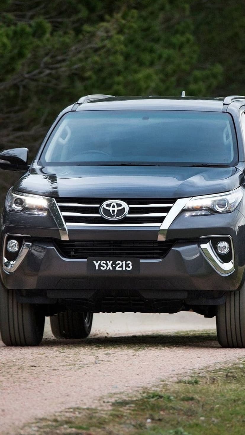 Dark grey Toyota Fortuner SUV with chrome accents on a dirt road surrounded by lush green forest trees.