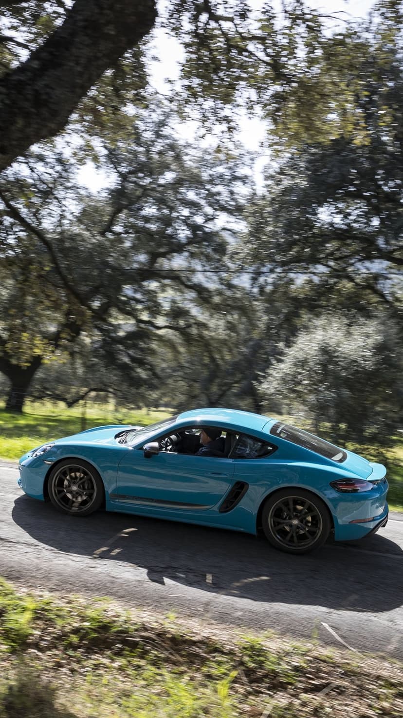 Blue Porsche 718 Cayman T driving fast on a forest road with motion blurred trees and sun-dappled lighting.