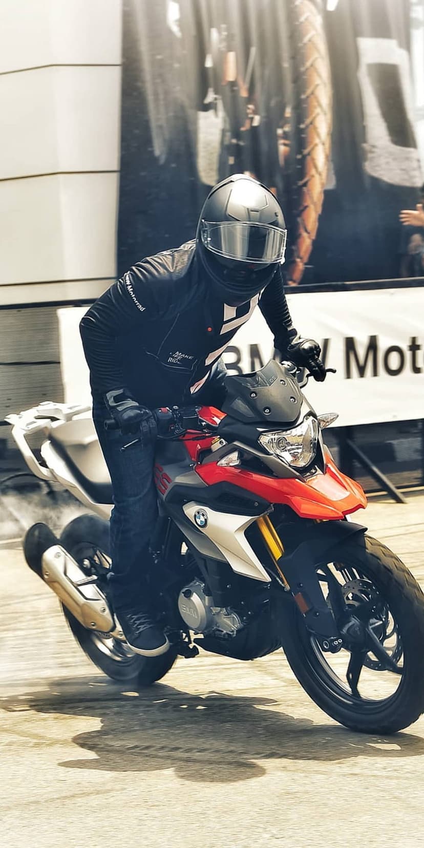 Motorcyclist in black gear leans a red and white BMW bike into a turn while rear tire smoke kicks up dust.