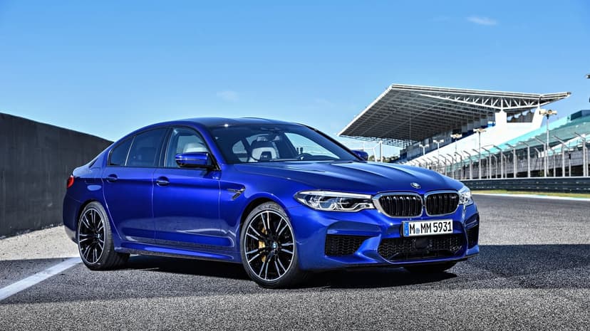 Blue BMW M5 sports sedan parked on a sunny racetrack with a grandstand background under a clear sky.