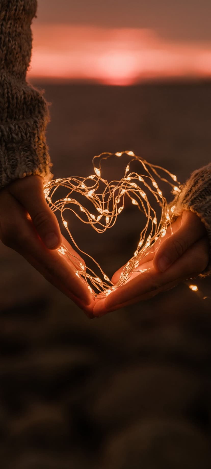 Hands forming a heart shape around glowing string lights against a blurred orange and pink sunset sky at dusk.