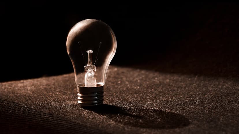 Clear incandescent bulb on a textured dark surface with a long shadow highlighting the glass and thin filament.