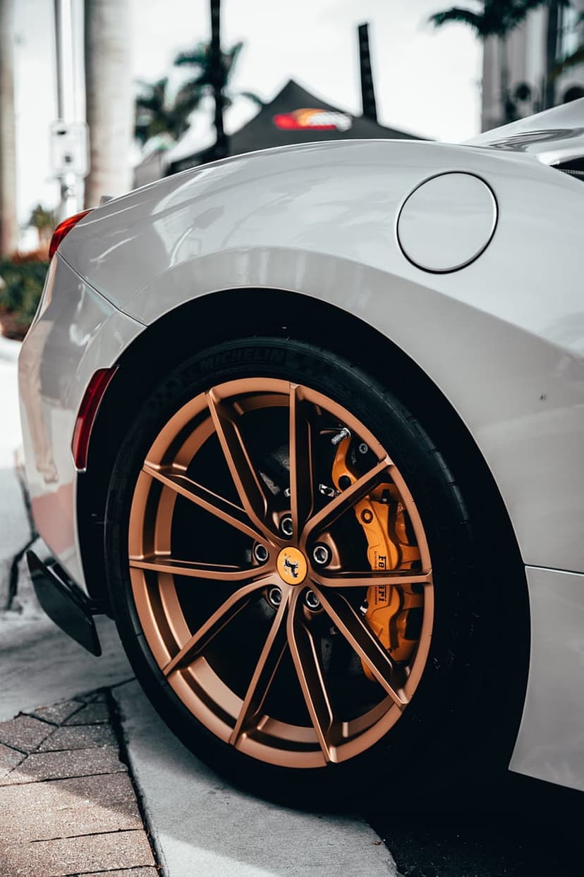 White Ferrari rear wheel featuring bronze multi-spoke alloy rims and bright orange brake calipers against palm trees.