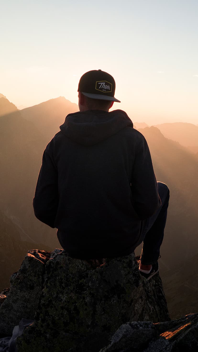 Person in a hoodie and cap sits on a rock overlooking misty golden mountain ridges at dawn in a vertical view.