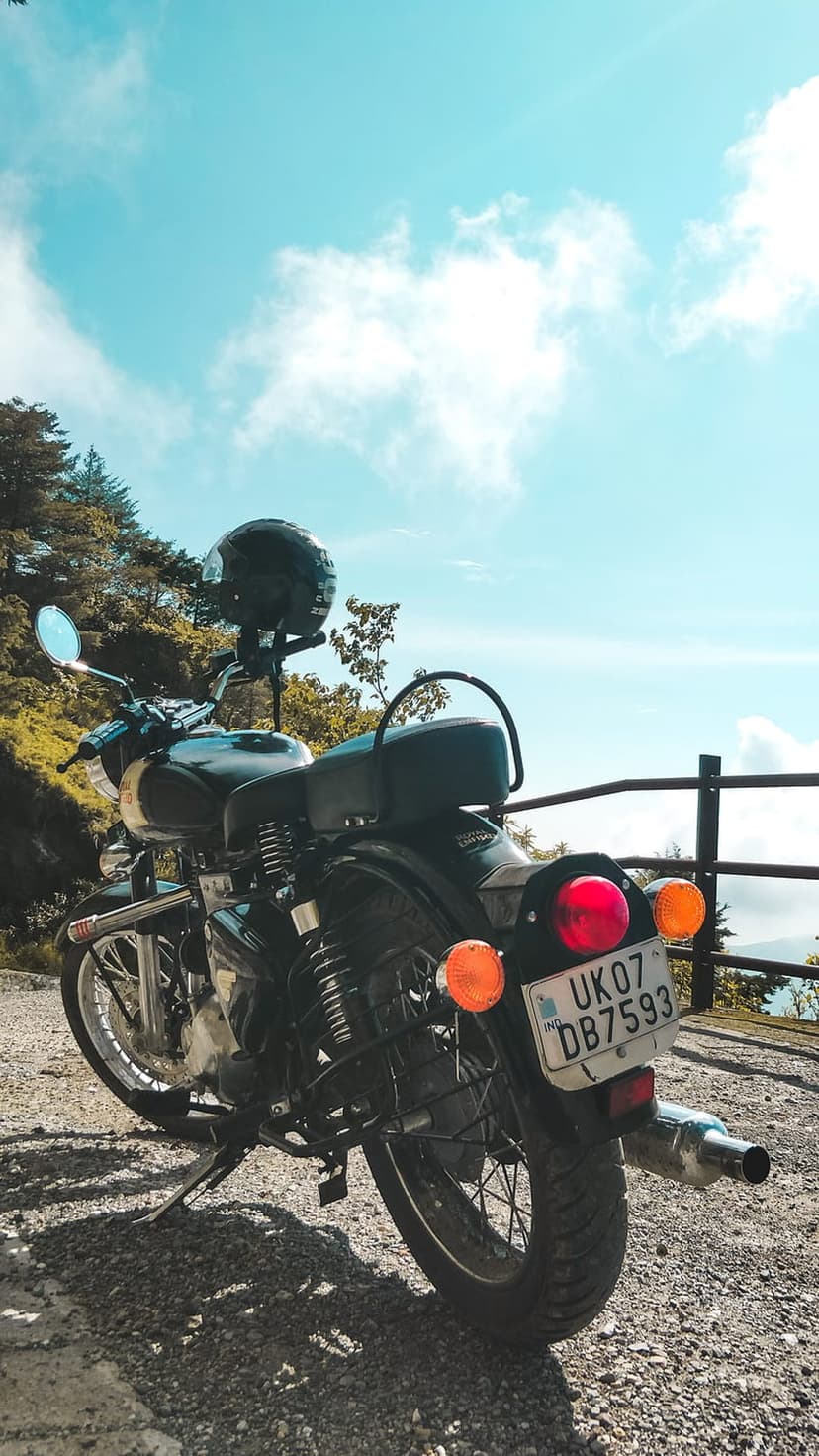 Black motorcycle with a helmet on the handlebars sits on a gravel hilltop overlooking a green forest and blue sky.