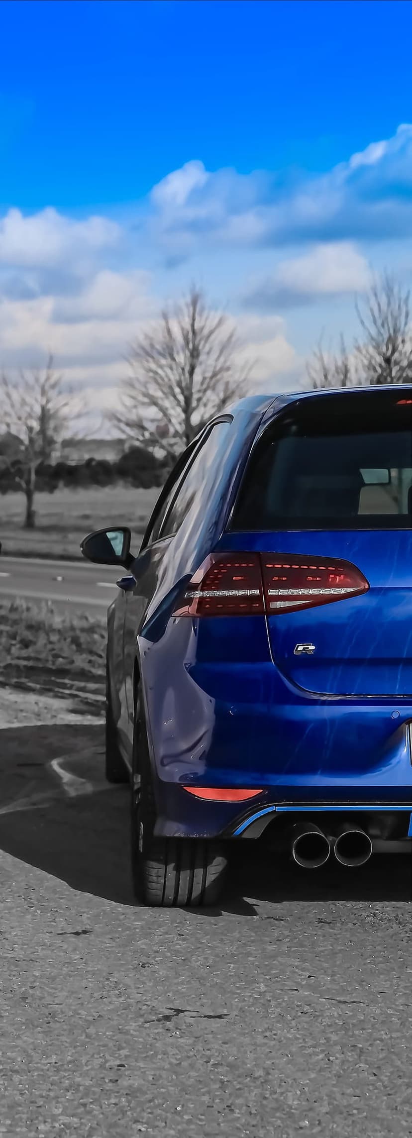 Blue Volkswagen Golf R tail lights and quad exhausts parked on asphalt under a clear blue sky and trees.