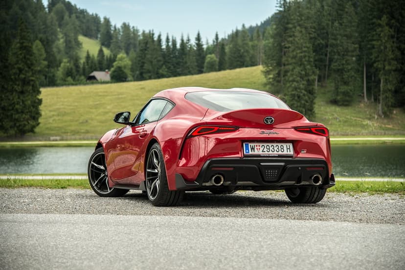 Rear view of a red Toyota GR Supra parked on a gravel road by a mountain lake and dense pine forest.
