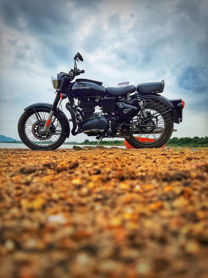 Black Royal Enfield motorcycle parked on a sandy beach under a cloudy sky with calm lake water in the background.