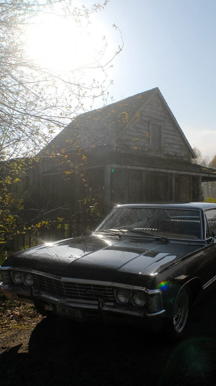 Black vintage Chevrolet Impala parked in front of a weathered wooden barn with sunny lens flares and shadows.