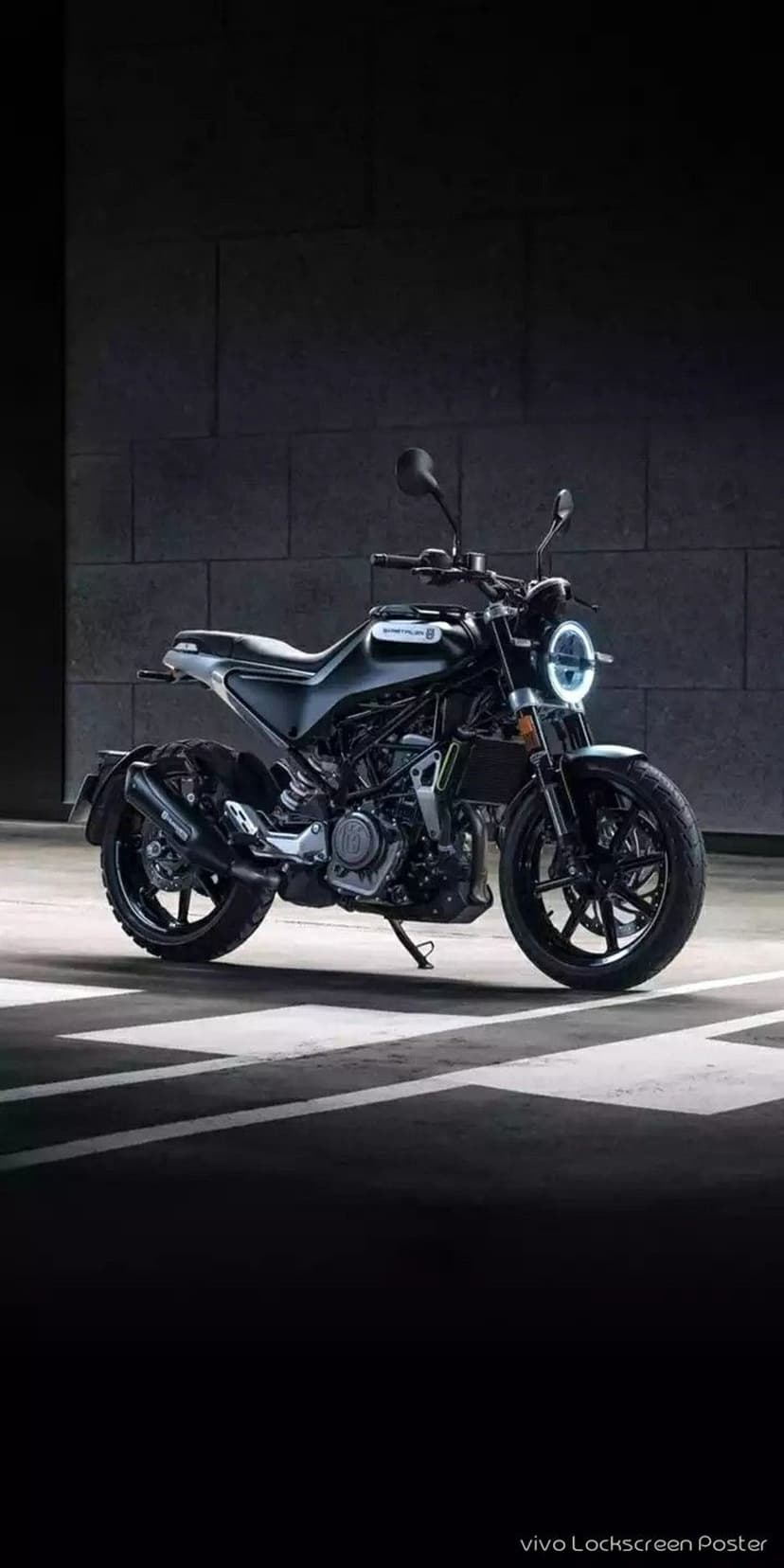 Husqvarna motorcycle with a round LED headlight parked on asphalt between white lines against a dark brick wall.