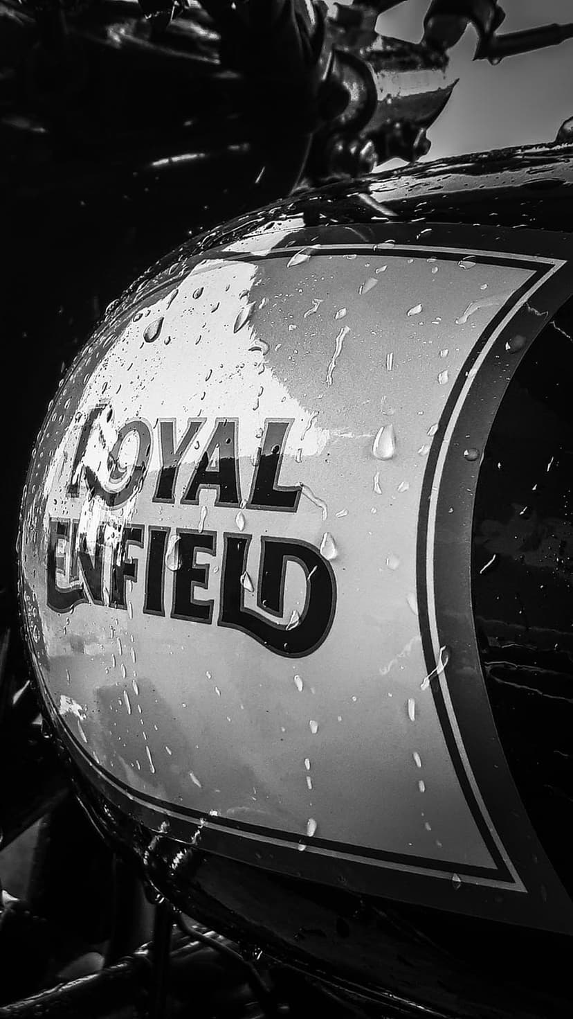 Royal Enfield motorcycle fuel tank with raindrops and metallic logo in a high-contrast black and white photograph.