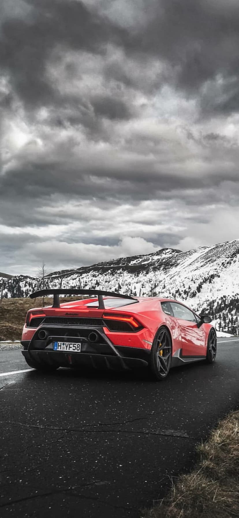 Red Lamborghini Huracan supercar on a wet mountain road with snow-covered peaks under a cloudy grey sky.